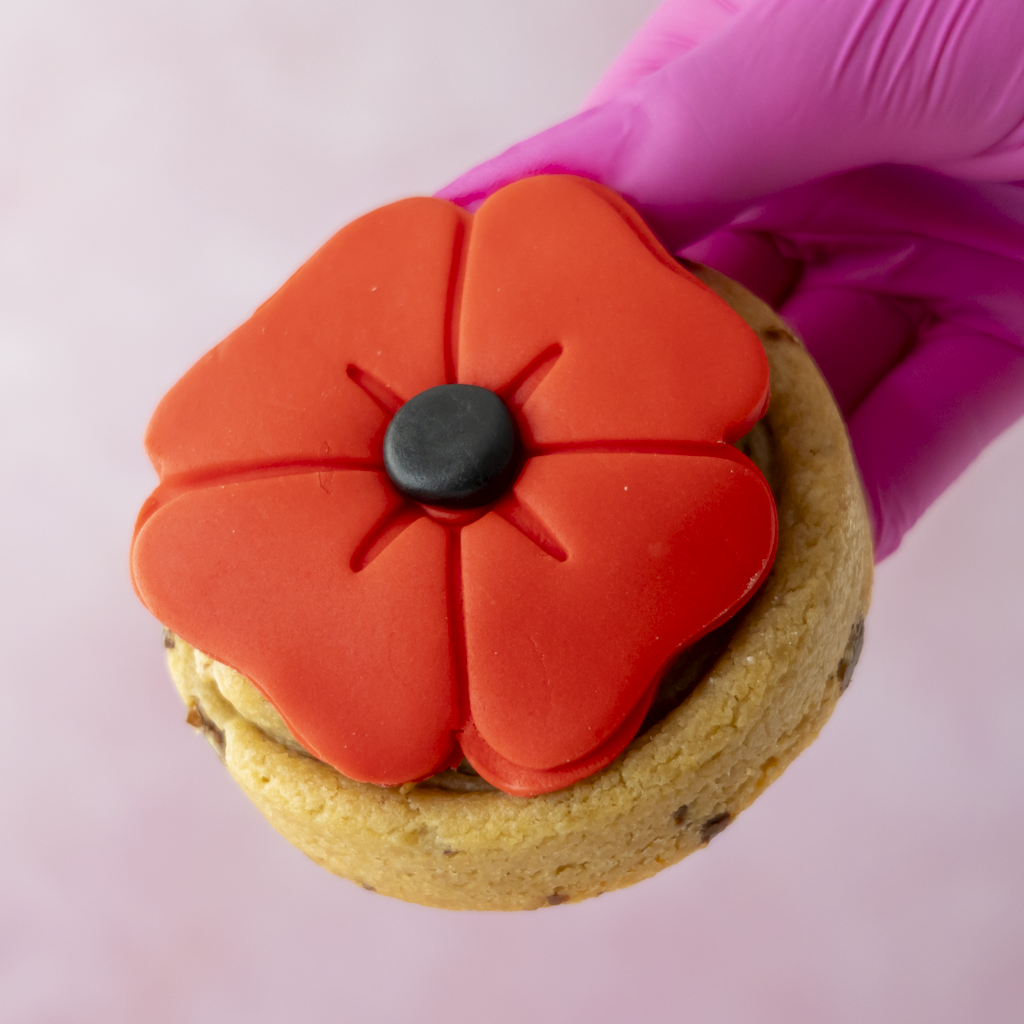 A close-up of a decorated cookie topped with a bright red poppy-shaped fondant decoration and a small black center, held by a hand wearing a pink glove against a soft pink background.
