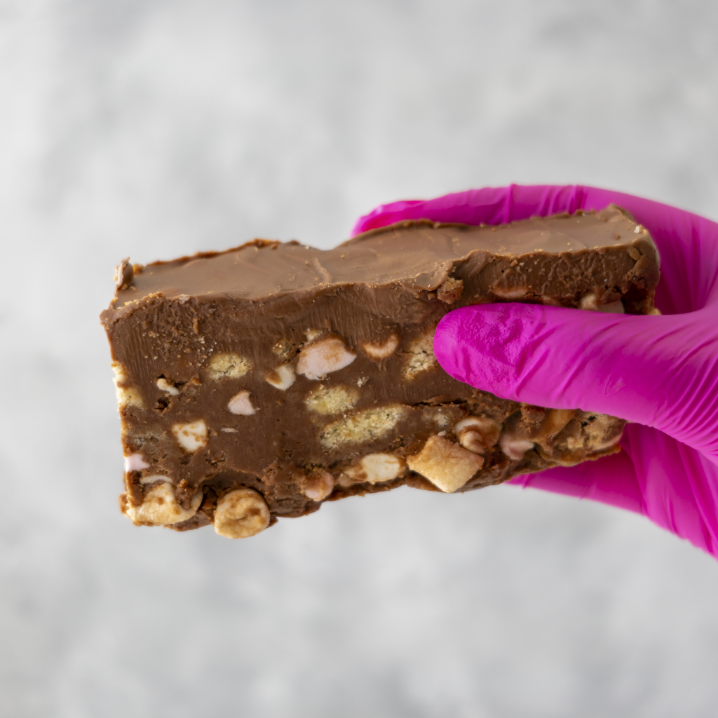 Rocky Mountain Rocky Road Slab fudge with nuts held by a hand wearing a pink glove on a light gray background