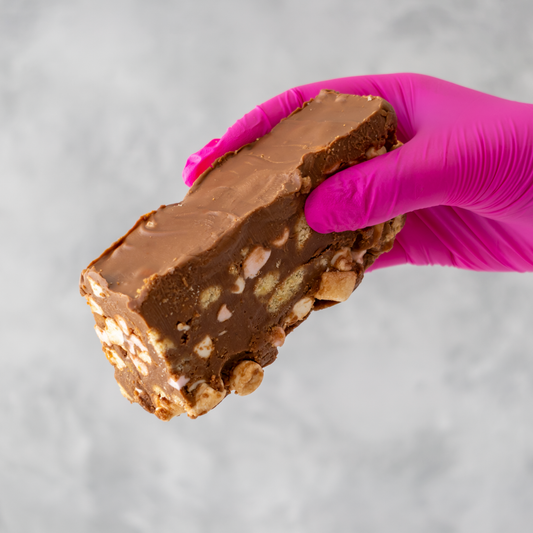 Handmade Rocky Mountain Rocky Road Slab with nuts held by a hand wearing a pink glove on a gray background