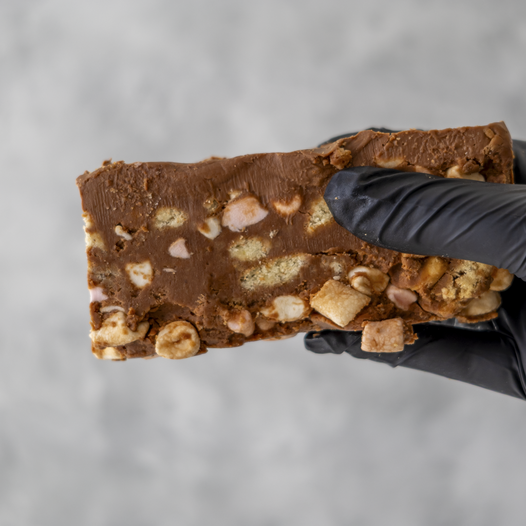 Hand holding a Handmade Rocky Mountain Rocky Road Slab with nuts and marshmallows against a gray background