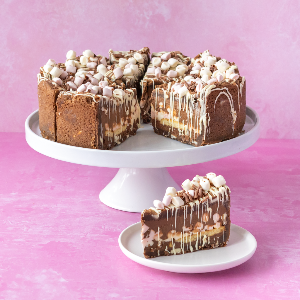 S’mores Cookie Pie with white chocolate drizzle and mini marshmallows, shown on a white cake stand with a slice on a small plate against a pink background.
