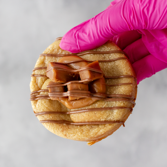 Sea Salt & Caramel Stuffed Deluxe Cookie with chocolate drizzle and caramel pieces held by a gloved hand on a light background