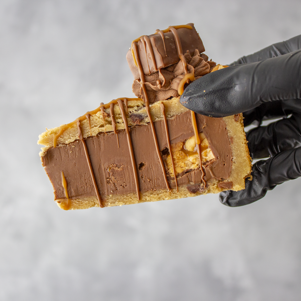 Cookie Pie Slice with Snickers held in black glove with chunky chocolate bar topping