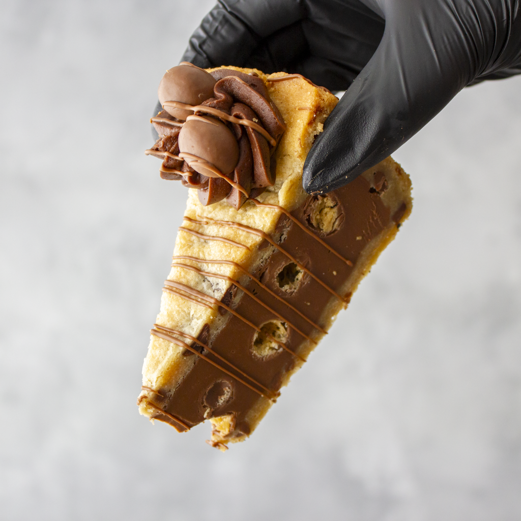 Hand holding a slice of dessert with chocolate drizzle anh chocolate balls against a gray background
