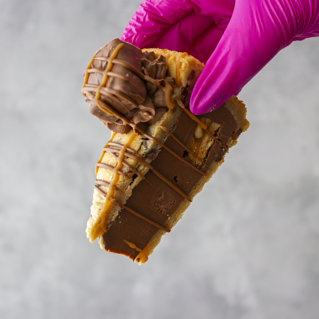 DoughGirl Cookie Pie Slice with Snickers held in pink glove on grey background