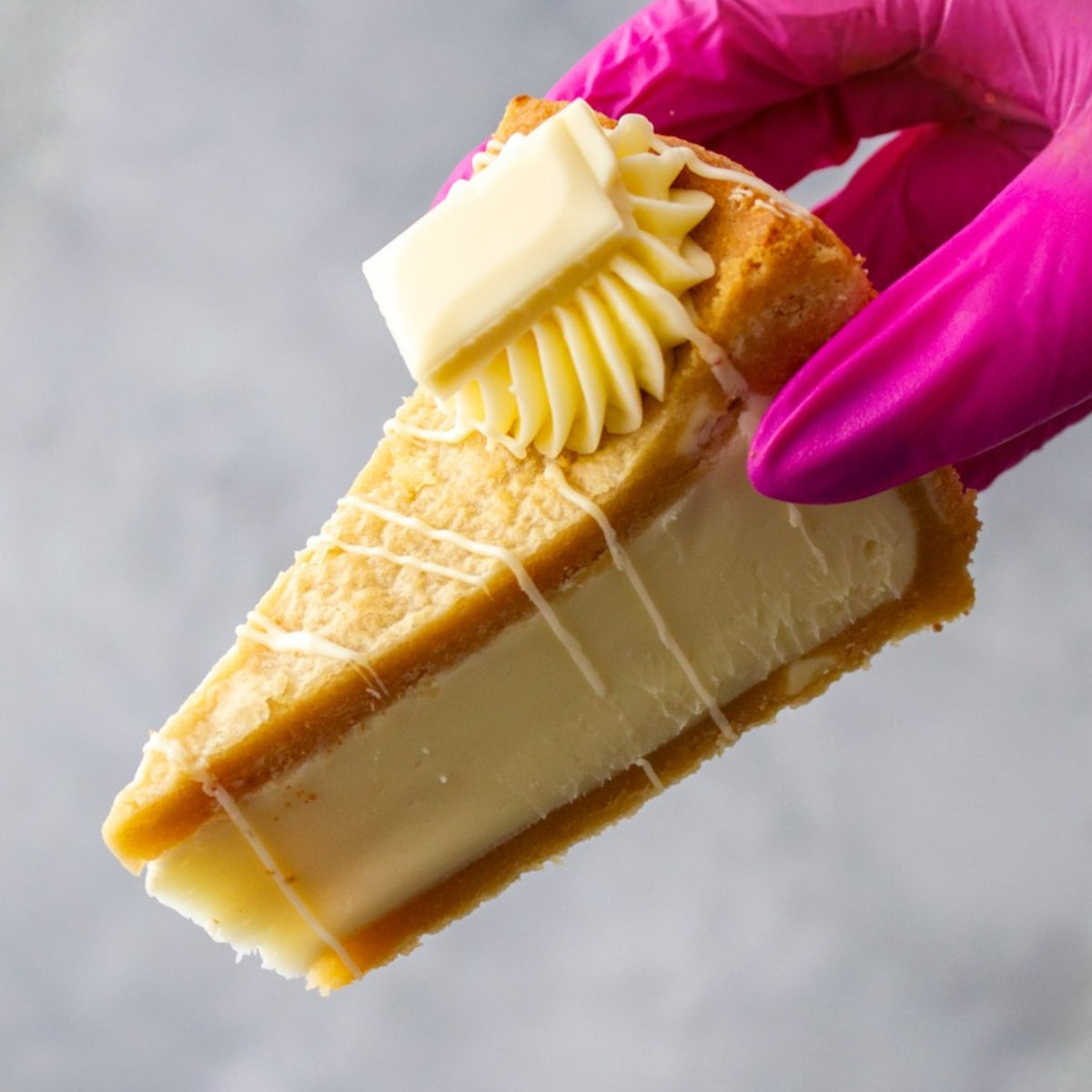 A close-up of a slice of Cookie Pie with Milkybar held by a hand wearing a bright pink glove, topped with a piped swirl of cream and a square of butter on a light grey background.