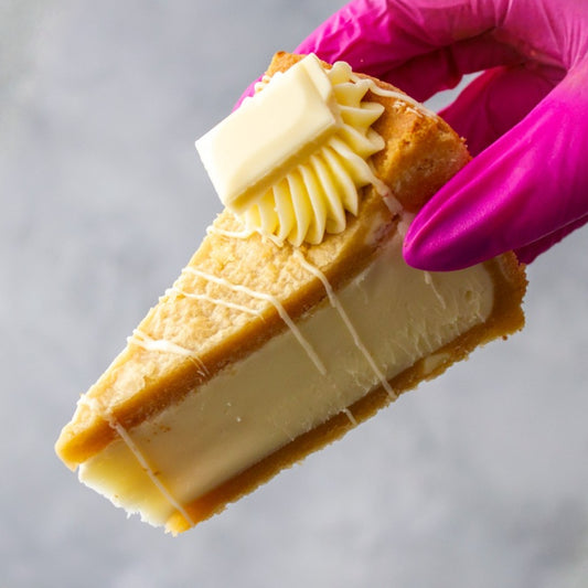 A close-up of a slice of Cookie Pie with Milkybar held by a hand wearing a bright pink glove, topped with a piped swirl of cream and a square of butter on a light grey background.