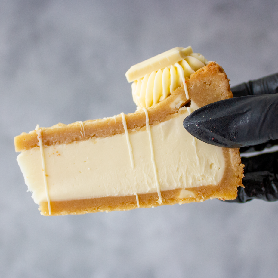 A close-up of a hand wearing a black glove holding a slice of cookie pie with a white chocolate filling, a golden-brown crust, and a drizzle on top, set against a soft grey background.
