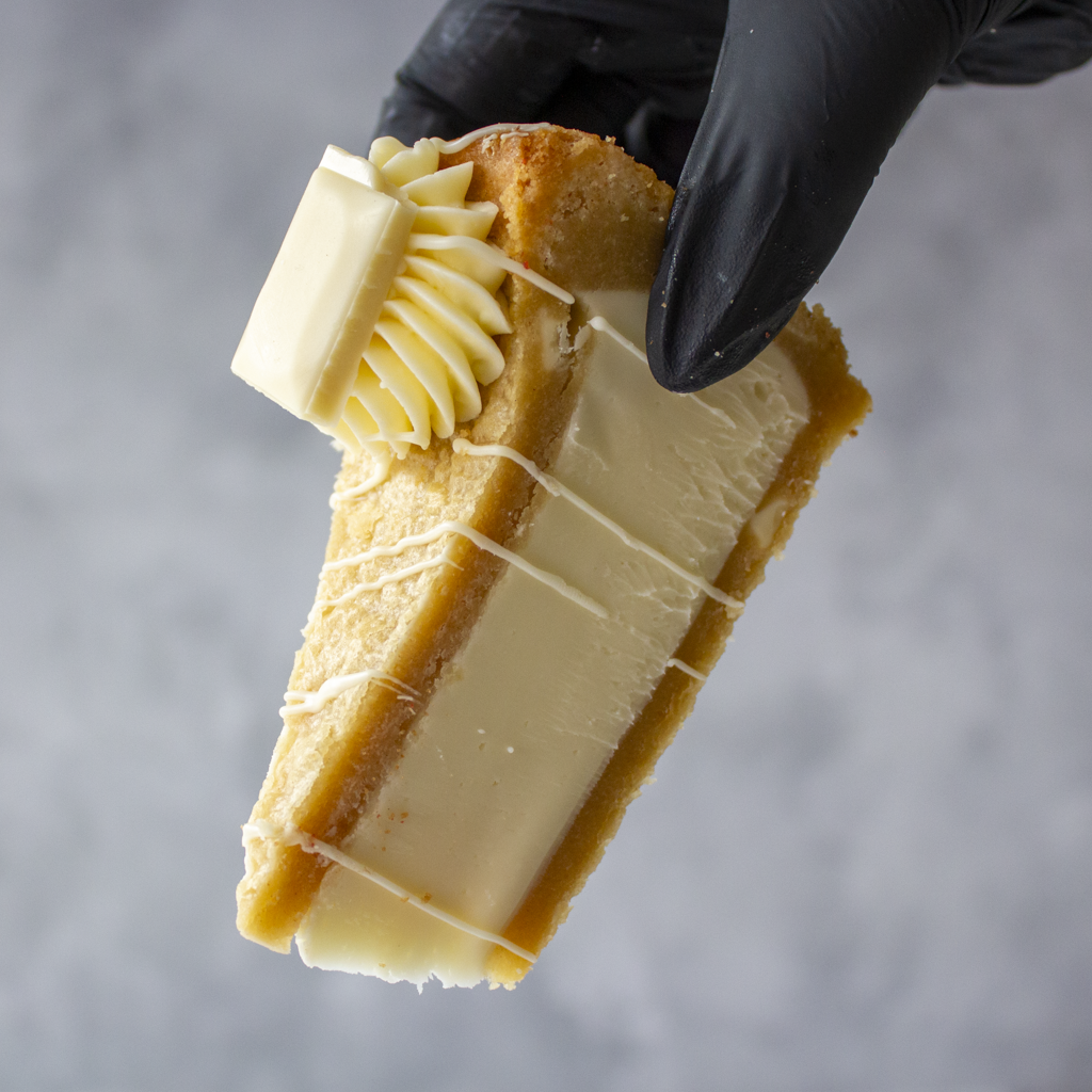 A close-up of a hand wearing a black glove holding a slice of Cookie Pie with Milkybar with white chocolate drizzle and a dollop of whipped cream, set against a neutral grey background.