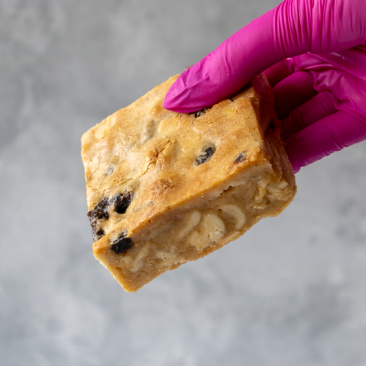 Hand holding a block of Sticky Toffee Blondie Slab wearing a purple glove on a gray background.