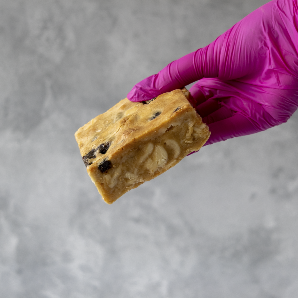 Hand wearing a purple glove holding a Sticky Toffee Blondie bar with chocolate chips against a gray background