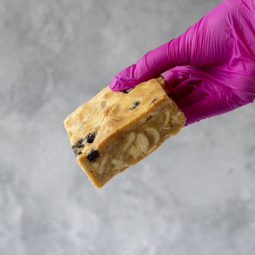 Hand wearing a purple glove holding a Sticky Toffee Blondie cookie bar with visible branding against a gray background