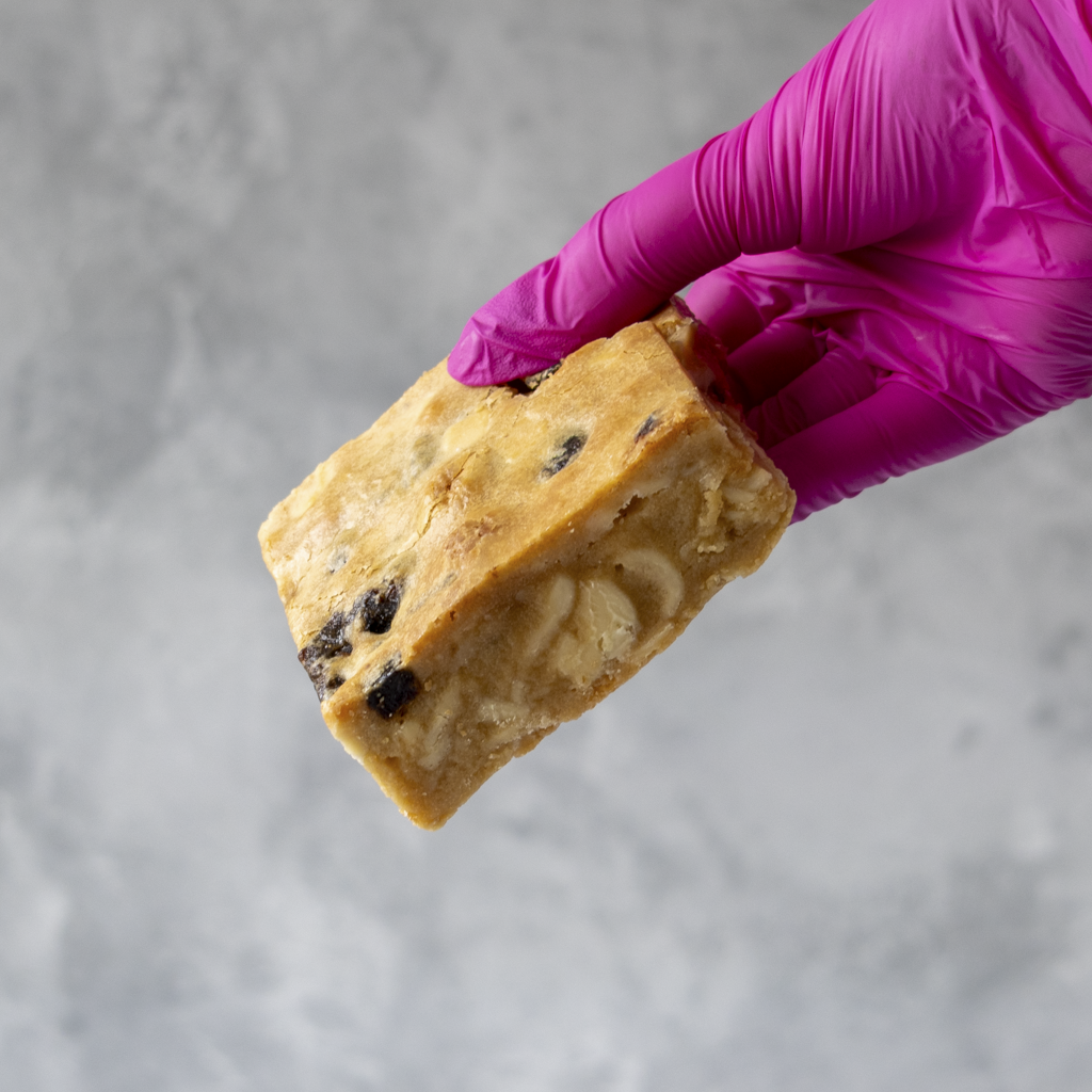Bar of Sticky Toffee Blondie with raisins held by a hand wearing a purple glove on a gray background