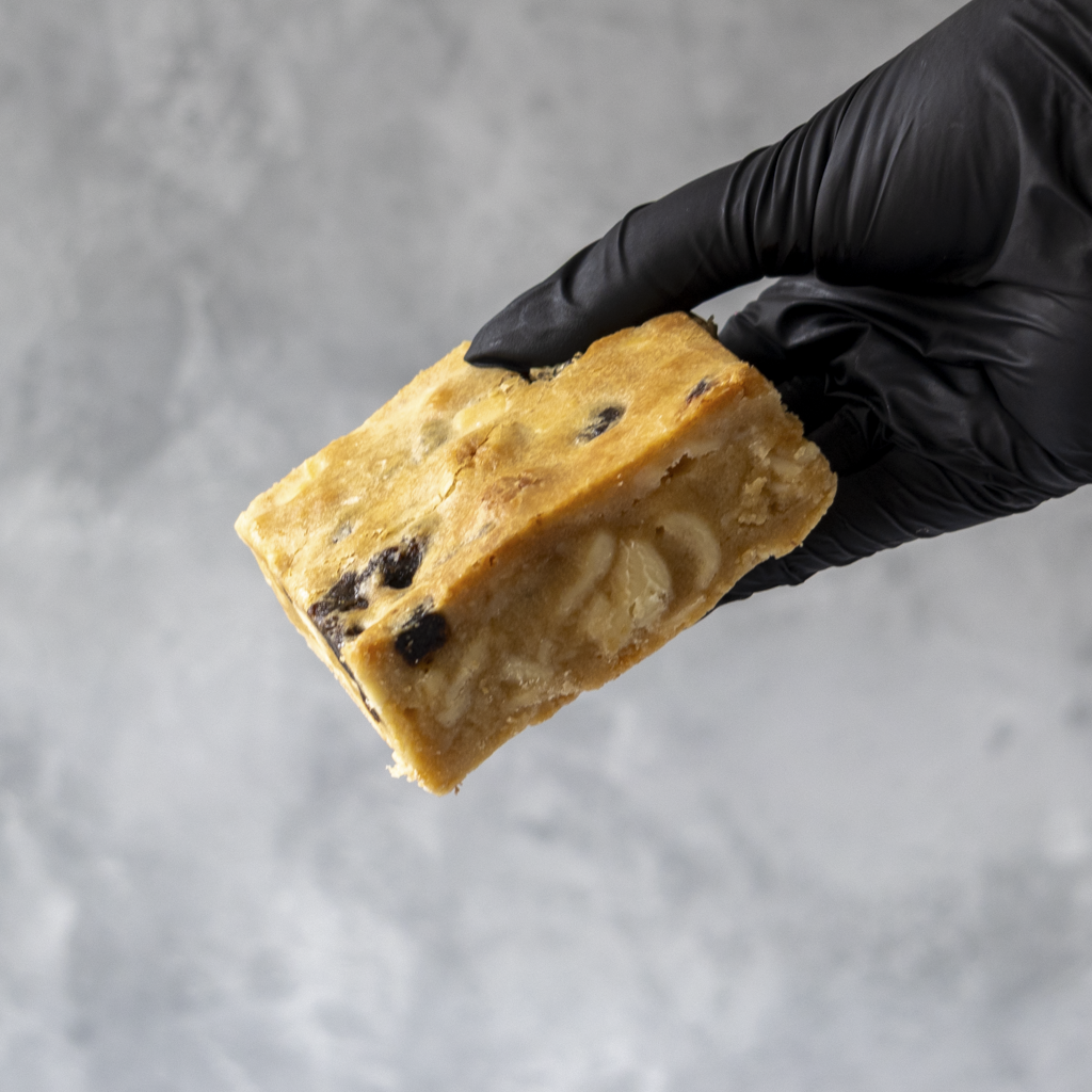 Hand wearing a black glove holding a piece of Sticky Toffee Blondie Slab against a gray background