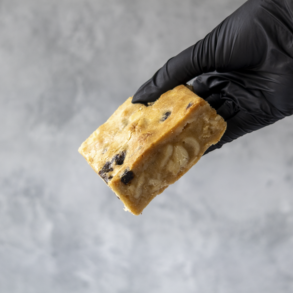 Hand wearing a black glove holding a Sticky Toffee Blondie Slab against a gray neutral background