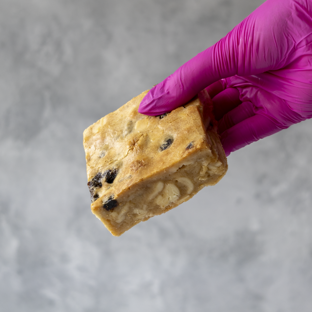 Hand wearing a purple glove holding a piece of Sticky Toffee Blondie Slab with chocolate chips against a gray background