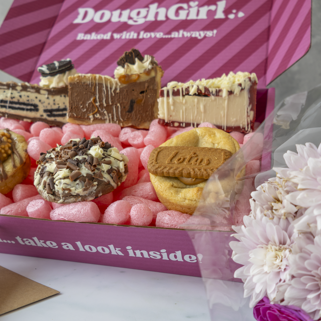 A pink gift box filled with assorted baked goods, including chocolate cookie pie slices, a cookie with a Lotus biscuit, and pink bubble wrap, with the box lid reading 'DoughGirl.