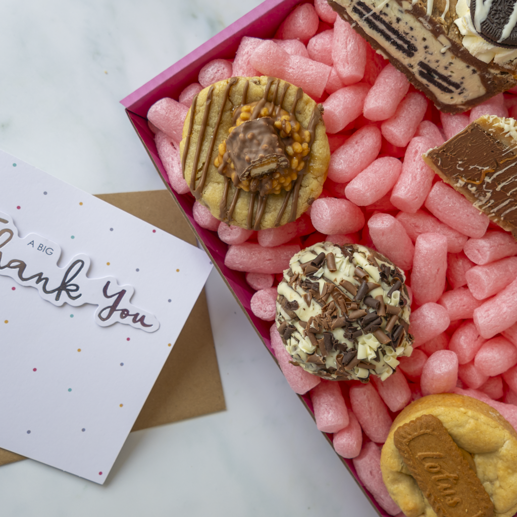 A gift box of assorted chocolate-covered cookies and biscuit pieces filled with pink packing foam, next to a white 'Thank You' card on a light marble surface.