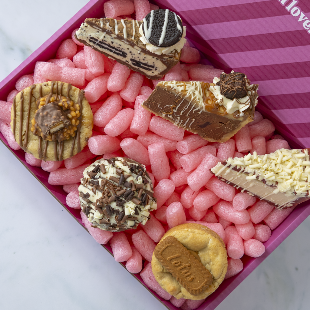 A boxed assortment of gourmet chocolate treats and cookies arranged in a pink gift box filled with pink packing peanuts. The assortment includes a chocolate sandwich cookie, a chocolate truffle with Oreo, a chocolate cake slice, a caramel and coconut dessert, a chocolate chip cookie, and a chocolate-covered biscuit.