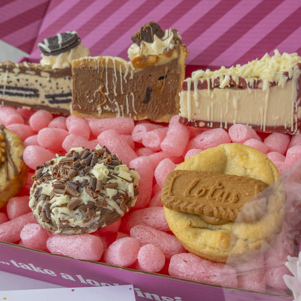 A close-up of assorted gourmet desserts arranged in a pink box. Visible treats include chocolate-dipped cookies, cream and chocolate drizzle cookie pie slices, a cookie with a biscuit bar, and a chocolate-and-cream swirl dessert.