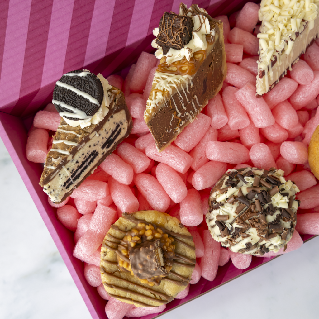 A close-up overhead view of assorted gourmet dessert pieces arranged in pink packing cases inside a pink striped box. The desserts include chocolate cookie pie slices, a cookie cream pie, a caramel-topped slice, and chocolate-covered cookies.