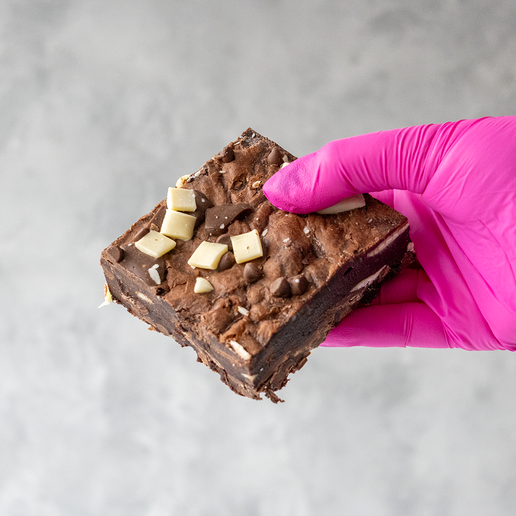 Triple Chocolate Brownie Slab by DoughGirl UK with milk, dark & white chocolate held by a hand in a pink glove on a gray background. Perfect for sharing or gifting