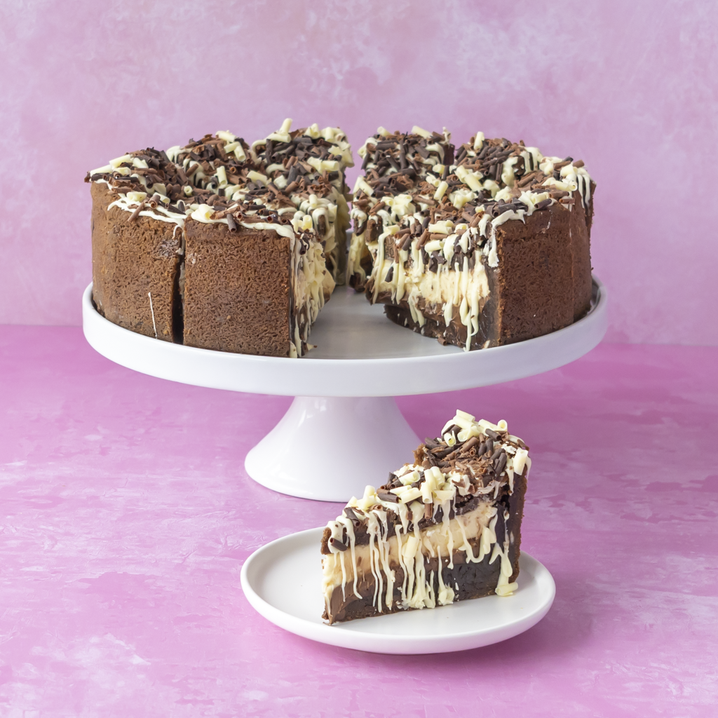 Chocolate cake with white chocolate drizzle and chocolate shavings, served on a white cake stand with a single slice on a small plate against a pink background.