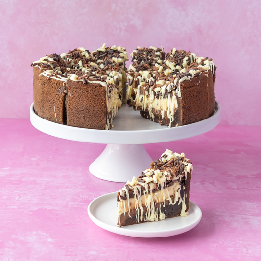 Chocolate cake with white chocolate drizzle and chocolate shavings, served on a white cake stand with a single slice on a small plate against a pink background.