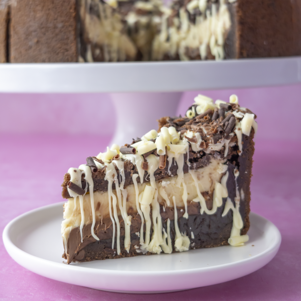 A slice of layered chocolate cookie pie with cream and white chocolate drizzle sits on a small white plate in front of a whole cake on a stand, against a pink background.