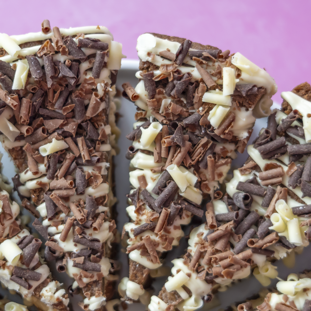 Close-up of chocolate bark pieces topped with white chocolate drizzle, chocolate shavings, and curled chocolate pieces, arranged on a plate against a pink background.
