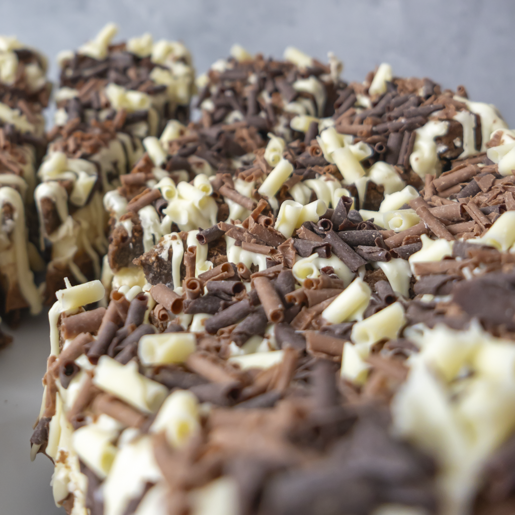 Close-up of a chocolate-covered treat or cookie topped with white chocolate shavings and dark chocolate curls, with more pieces blurred in the background.