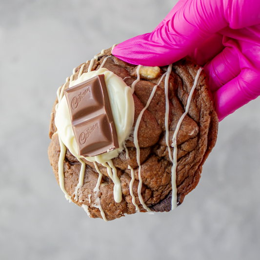 Triple Chocolate Stuffed Deluxe Cookie with chocolate drizzle and Cadbury bar held by a hand in a pink glove on a gray background