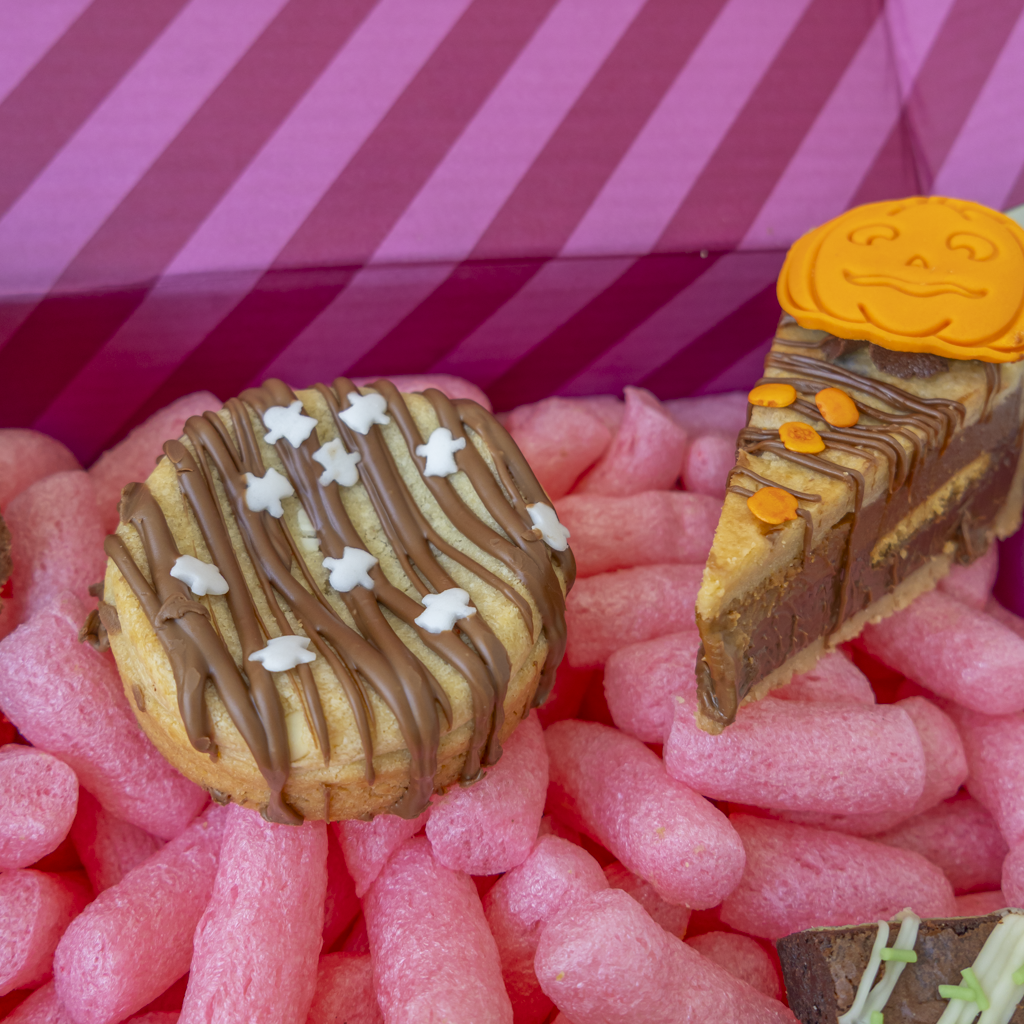 Two Halloween-themed gourmet treats presented in a pink box: a round chocolate-drizzled cookie with white ghost sprinkles and a triangular slice of layered cookie pie topped with an orange pumpkin face decoration.