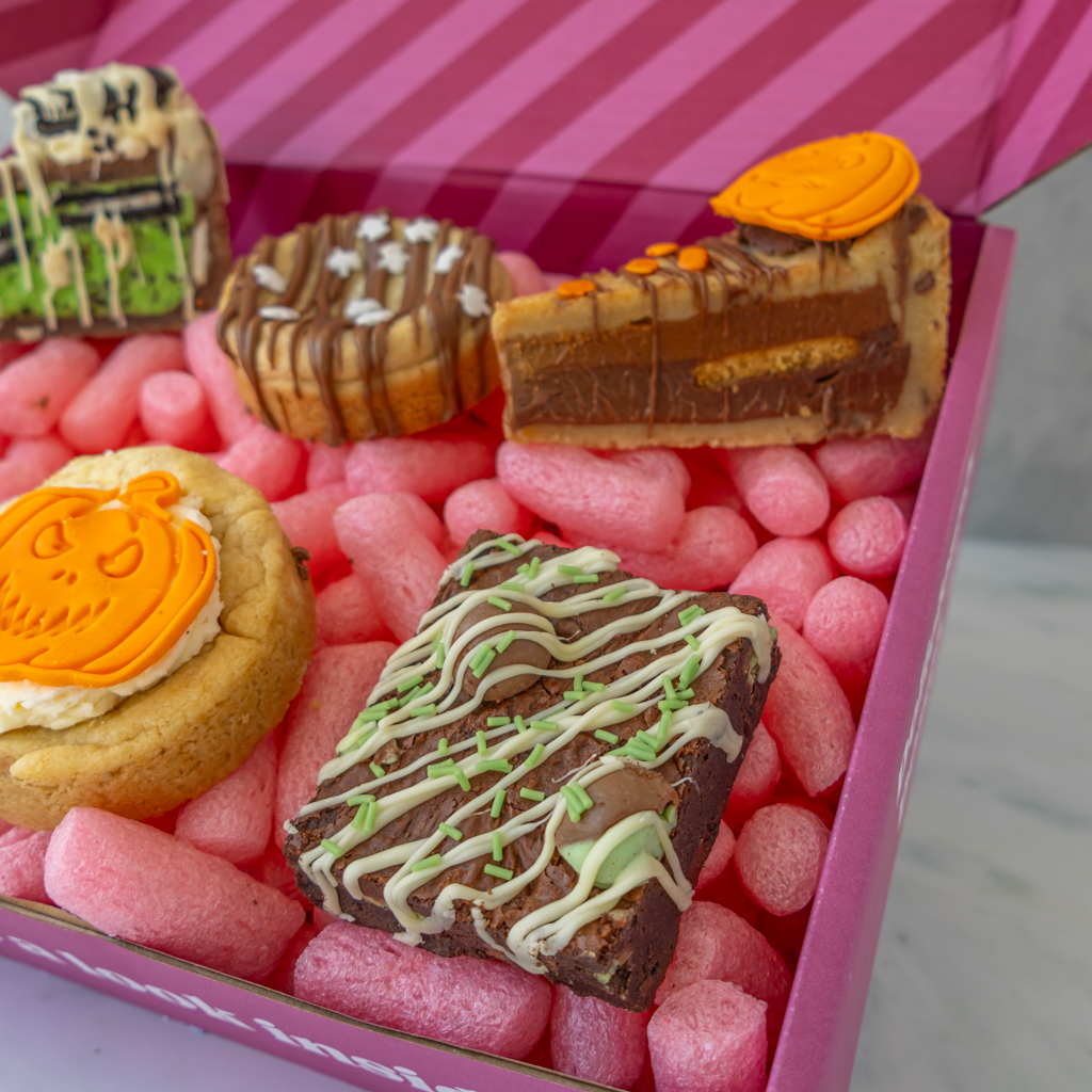 A close-up of assorted Halloween-themed gourmet treats arranged in pink packing foam inside a pink box. The treats include a pumpkin-faced cookie, a chocolate chip cookie with orange icing, a slice of chocolate cookie pie with orange topping, a cookie with drizzle, and a brownie with green and white drizzle.