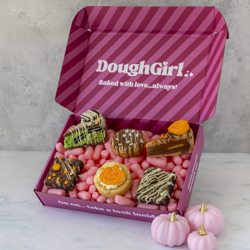 An open pink DoughGirl gift box filled with assorted baked Halloween-themed cookies, cookie pies, and slabs, with small pink pumpkins, photographed on a light marble surface against a grey wall.