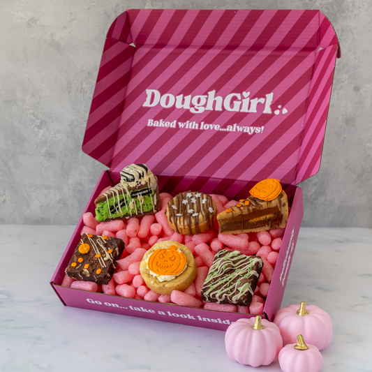 An open pink DoughGirl gift box filled with assorted baked Halloween-themed cookies, cookie pies, and slabs, with small pink pumpkins, photographed on a light marble surface against a grey wall.