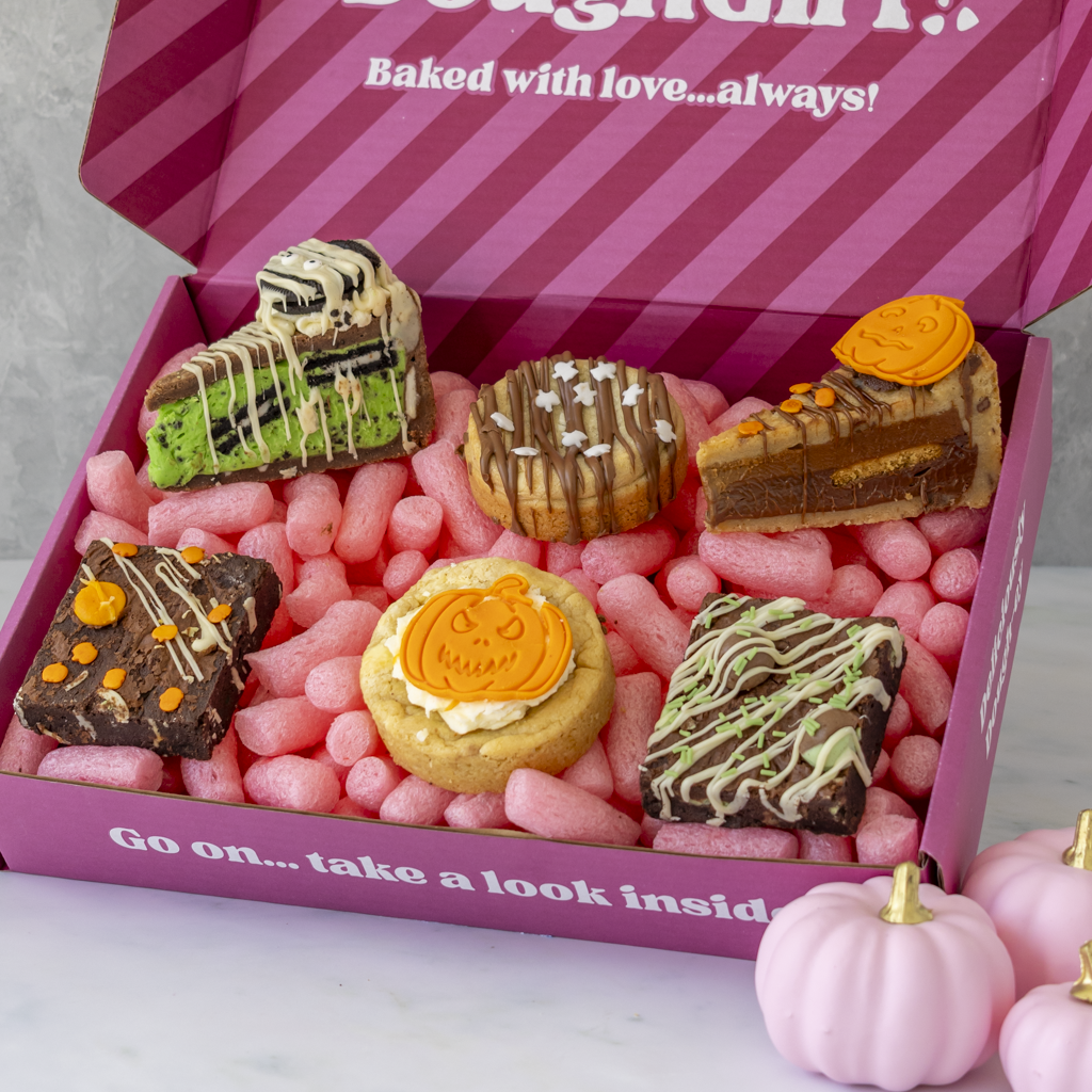 A pink gift box filled with assorted Halloween-themed baked goods, including cookies, brownies, and a slice of cake. The box lid reads 'Baked with love...always"