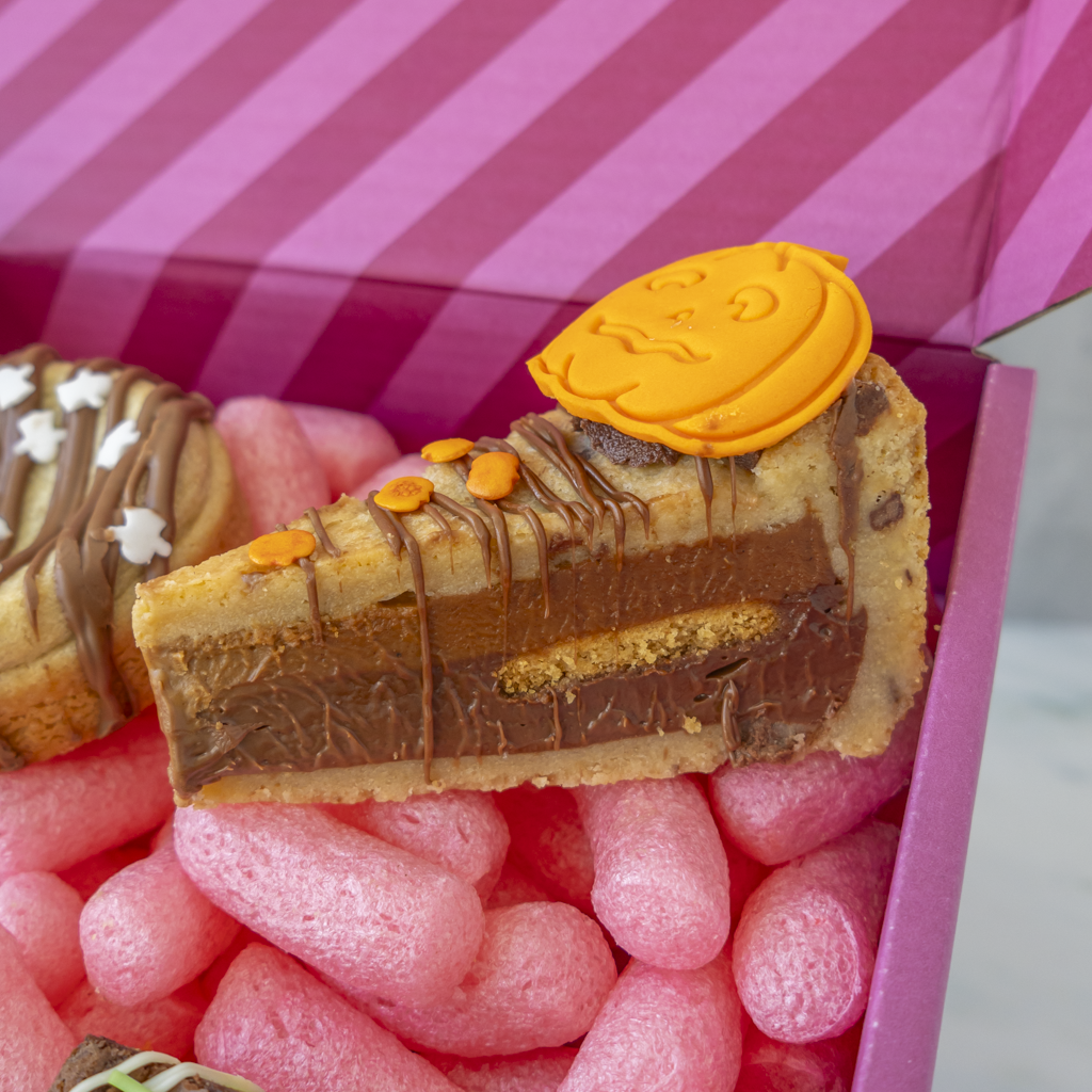 A slice of layered cookie pie with chocolate drizzle and a small orange pumpkin decoration, presented in a pink box alongside another pastry partially visible.