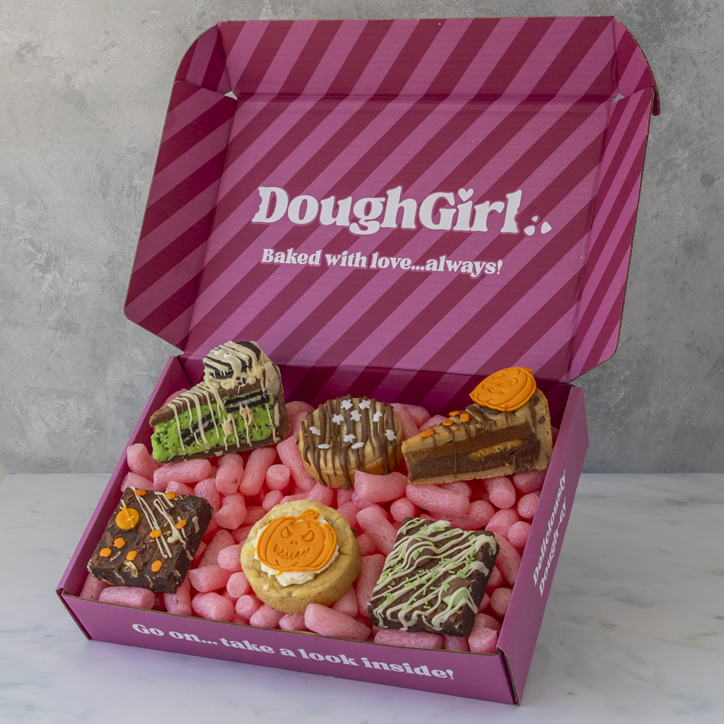An open pink DoughGirl gift box filled with assorted brownies, cookie pie slices, and cookie treats, set against a grey textured background.