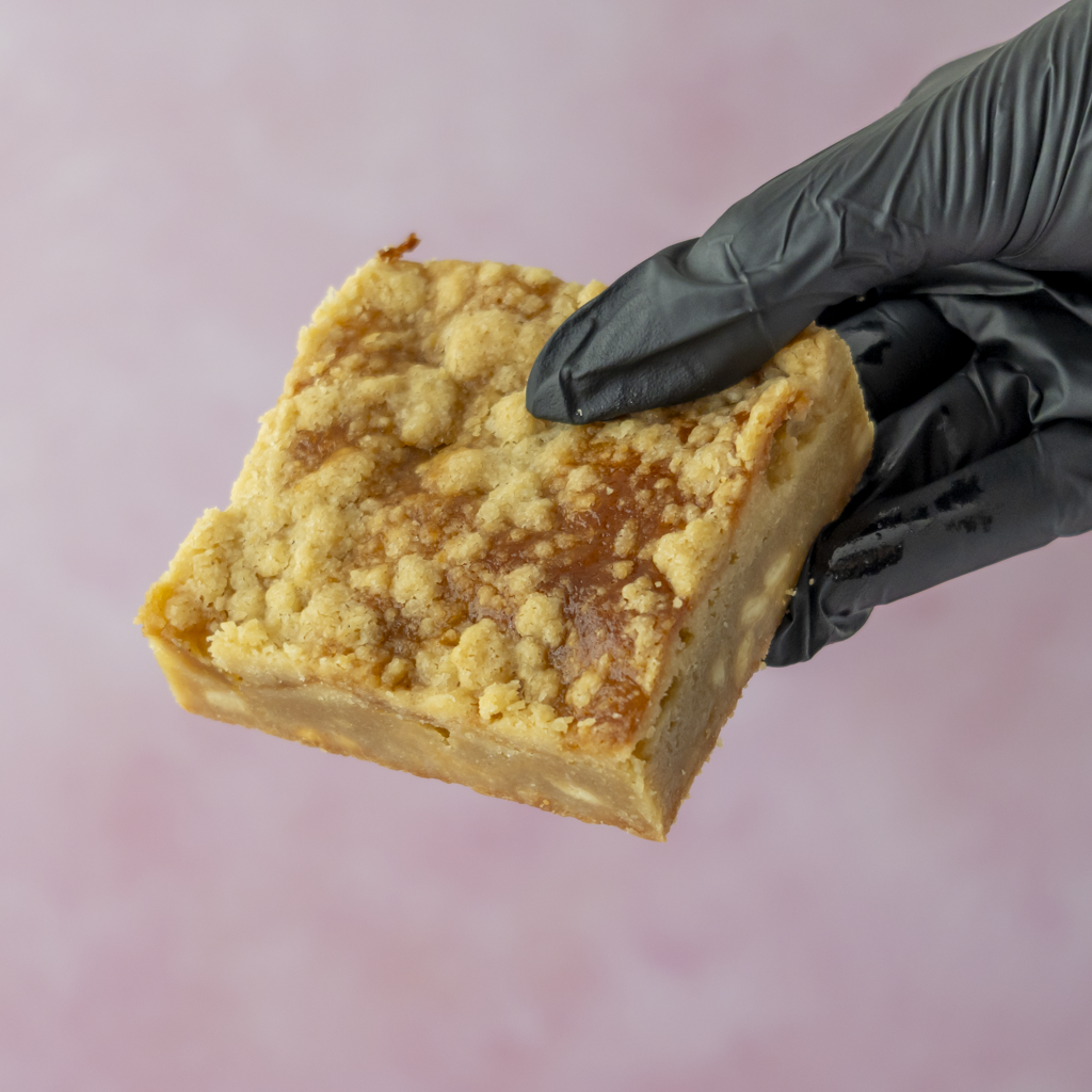 A hand wearing a black glove holds a square slice of Sticky Toffee Apple Crumble Blondie Slab against a soft pink background.