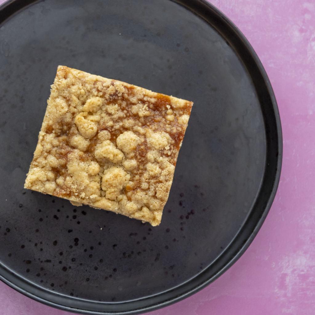 A single square crumb-topped blondie bar sits on a round black plate against a pink background.