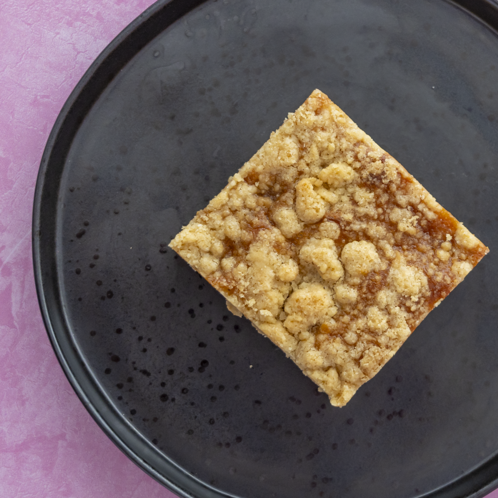 A single square slice of crumb-topped blondie slab placed on a round black plate against a pink background.