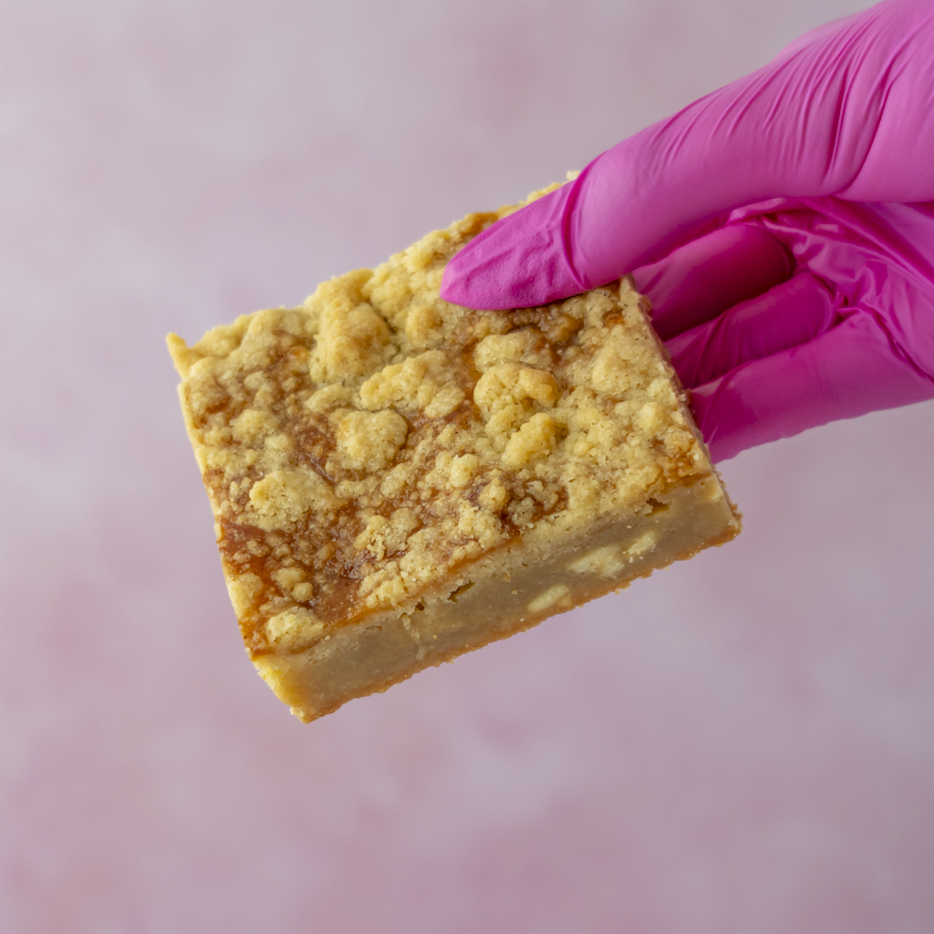 A square slice of golden-brown crumb-topped blondie slab held by a hand wearing a bright pink glove, set against a soft pink background.