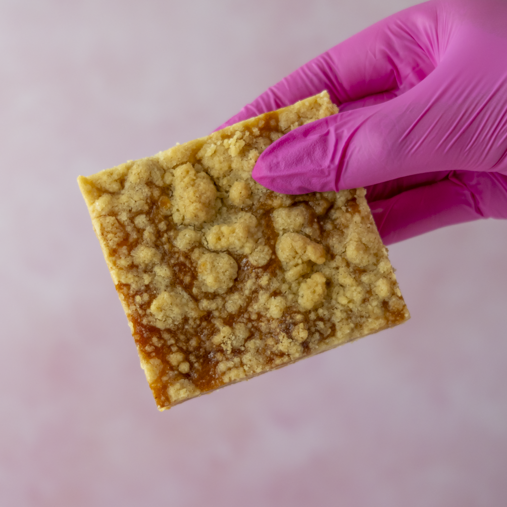 A hand wearing a bright pink glove holds a square Sticky Toffee Apple Crumble Blondie Slab with a glossy amber-orange filling against a soft pink background.