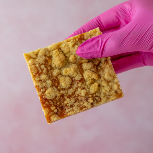 A hand wearing a bright pink glove holds a square Sticky Toffee Apple Crumble Blondie Slab with a glossy amber-orange filling against a soft pink background.