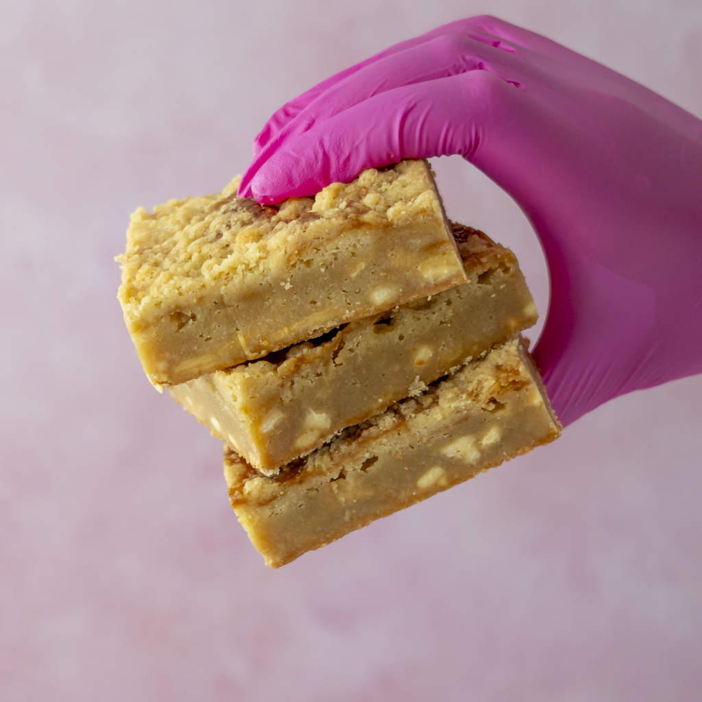 Three Sticky Toffee Apple Crumble Blondie Slabs stacked on a hand wearing a bright pink glove, set against a soft pink background.