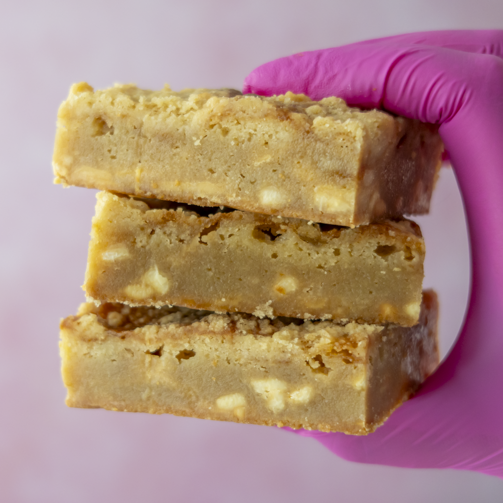 Three golden-brown baked blondie slabs stacked on a hand wearing a bright pink glove, set against a soft pink background.