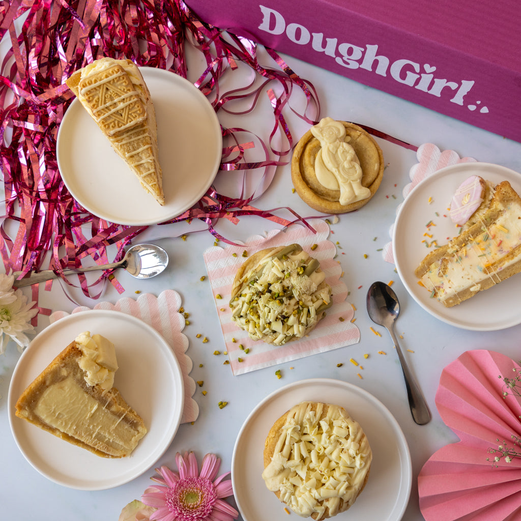 A styled flat-lay of several dessert slices and cookies arranged on white plates with pink and silver party decorations, including a pink box labeled DoughGirl.