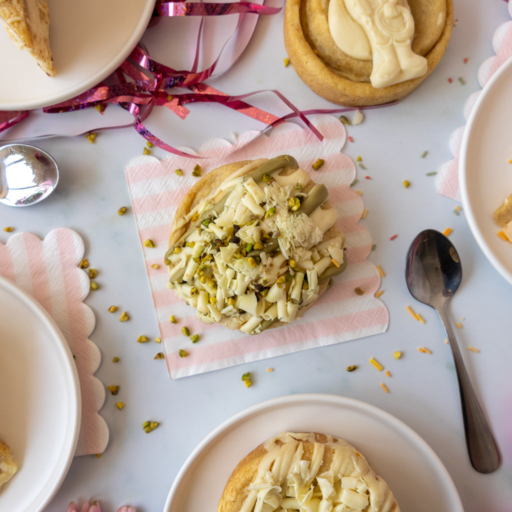 A top-down styled food scene showing a cookie topped with white chocolate and chopped pistachio on a pink-and-white striped napkin, surrounded by plates, spoons, ribbon, and other pastries on a light surface.