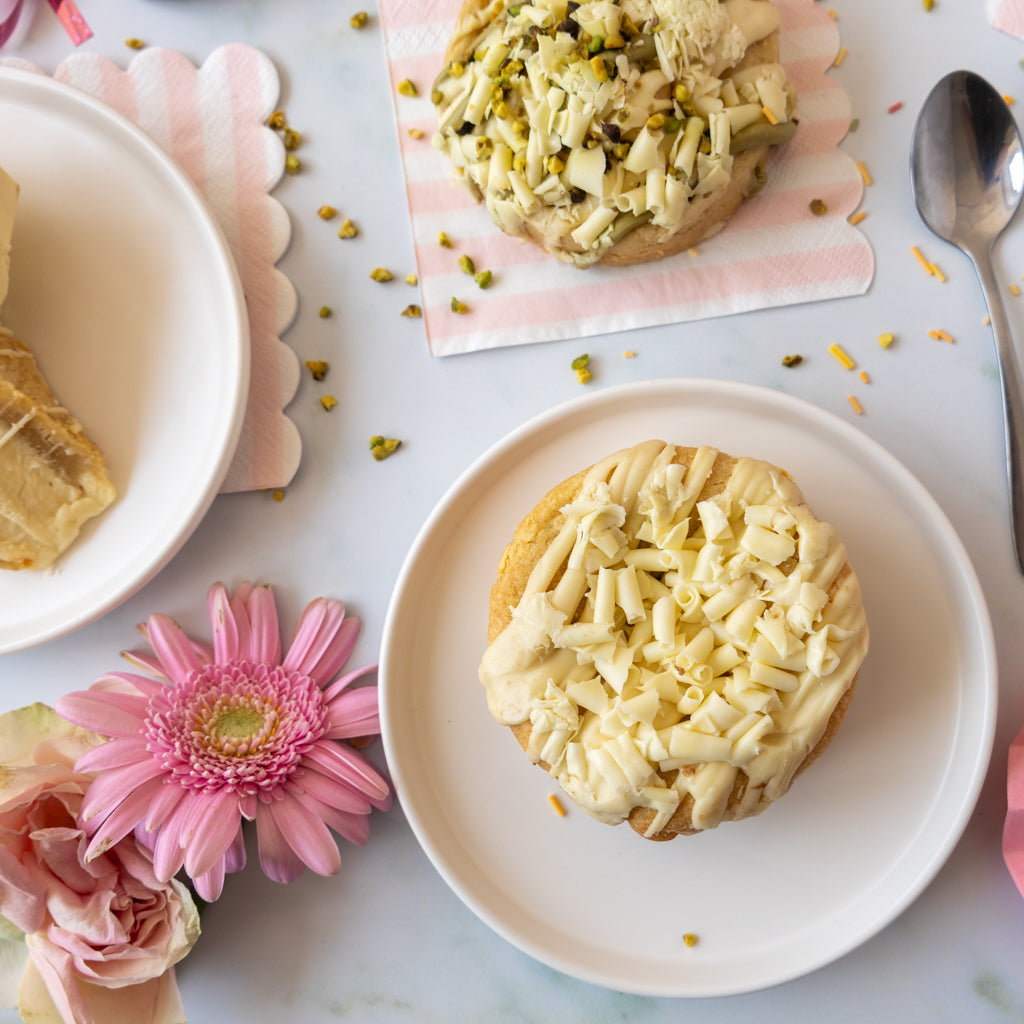 Two small cookies topped with white icing and chopped pistachio, arranged on white plates on a light marble surface with pink flowers, a spoon, and a pink-and-white napkin.